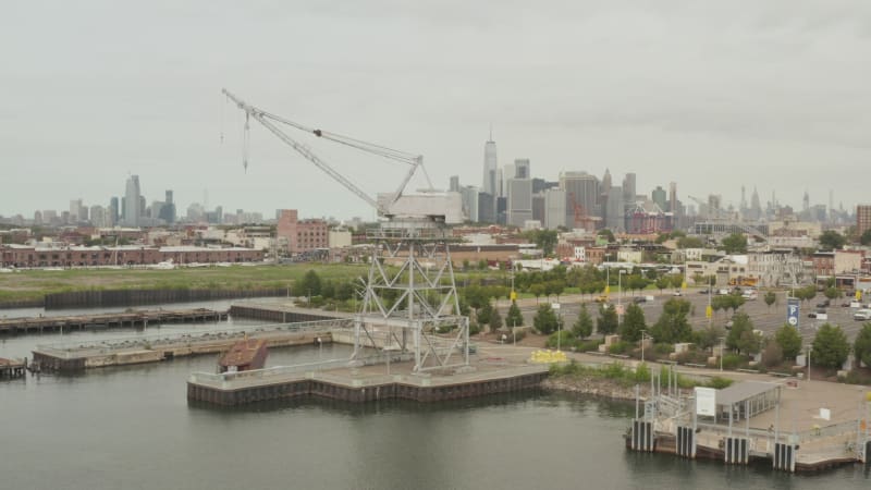 Flying in between industrial cranes in docks with New York City skyline in background and river