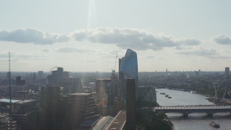 Slide and pan shot of tall modern One Blackfriars skyscraper on waterfront at bridges across water. View against sunshine. London, UK