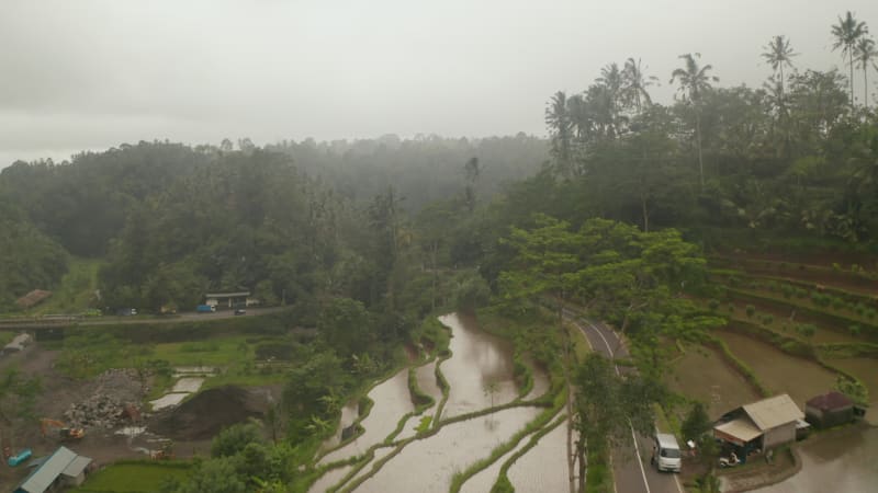 Cars driving on a winding road through rural terraced farm fields in Bali. Aerial dolly shot of rice fields full of water in rural Indonesia