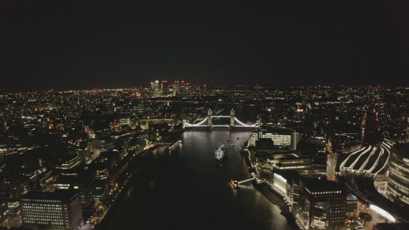 Backwards fly above Thames river. Aerial view of white colour illuminated Tower Bridge. Revealing of Shard skyscraper. London, UK