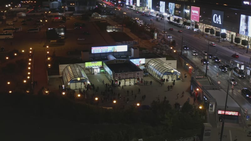 Slide and pan footage of illuminated Metro entrance near wide multilane street at night. People walking from subway. Warsaw, Poland