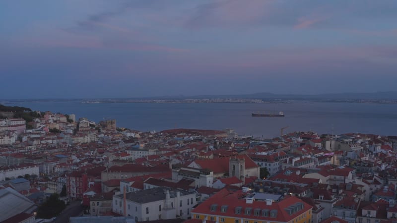 Aerial view of evening city rooftops. Drone flying over red roofs after sunset. Large water surface of Tegus river in background. Lisbon, capital of Portugal.