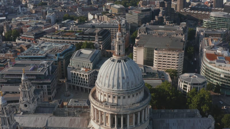 Fly over dome of historic Saint Pauls Cathedral in afternoon bright sun. Tilt down on decorated tower with cross on top. London, UK