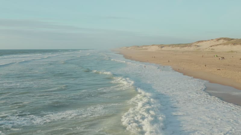 Waves Crashing onto Shore Line of Beautiful Beach in Afternoon light with People having fun, Aerial high angle circling slow