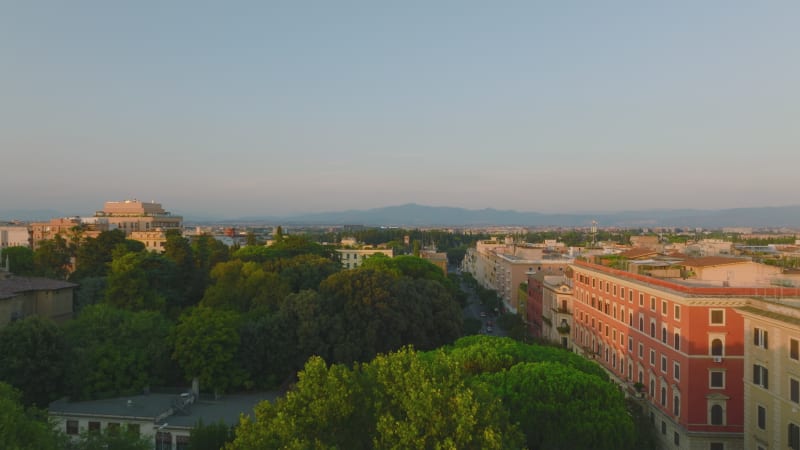 Aerial view of grown trees in park between blocks of apartment buildings lit by low sun. Forwards fly above urban borough. Rome, Italy