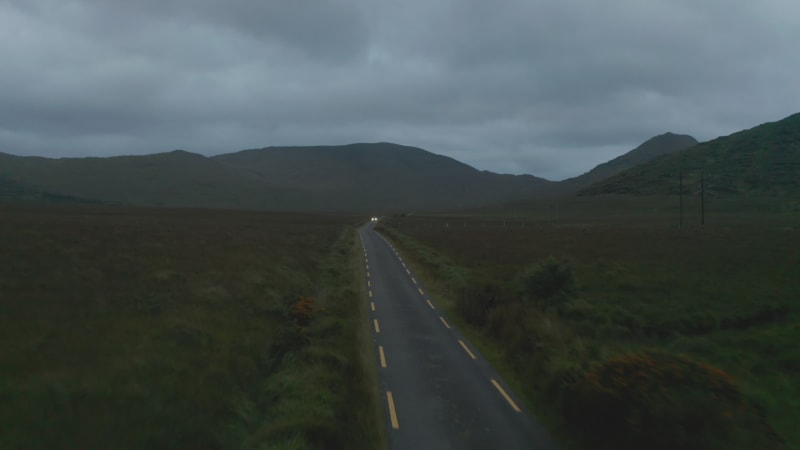 Forwards fly above narrow country and passing against car. Cloudy day at dusk. Mountain ridge in background. Ireland