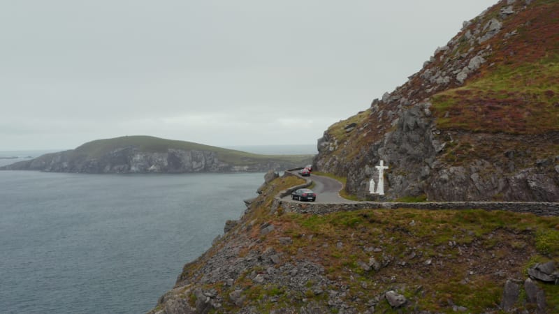 Car waiting in curve on oncoming vehicle. Difficult driving on panoramic coastal route. White sculpture near road, Cross at Slea Head. Ireland