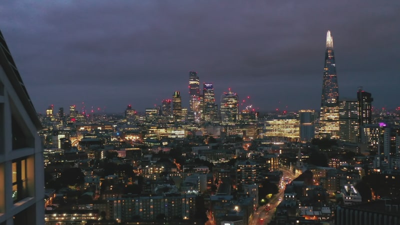 Panoramic aerial view of illuminated downtown skyscrapers at night. Backwards reveal of modern high rise apartment building. London, UK