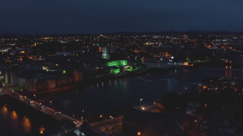 Fly above night city, green illuminated large building on Shannon river waterfront. Limerick, Ireland