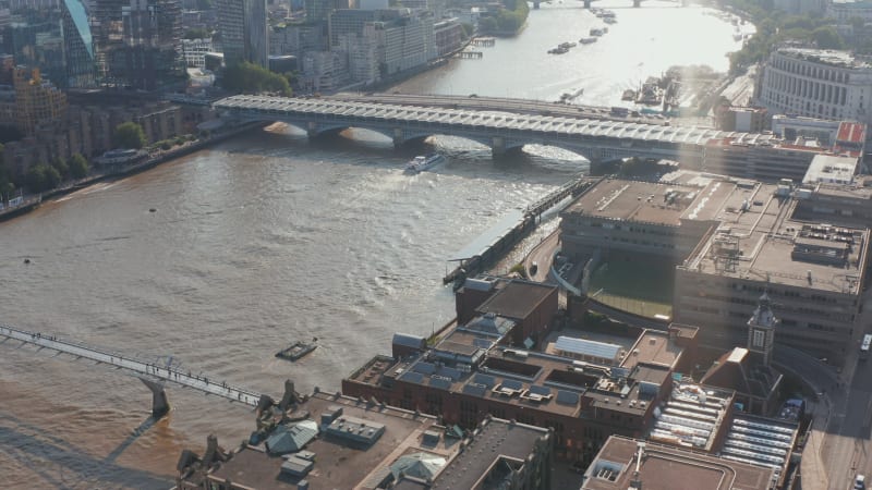 High angle view of buildings on waterfront at Millennium footbridge across River Thames. Tilt up reveal of turning river and modern buildings on south bank. View against sunshine. London, UK