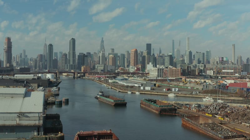 aerial descending footage of ship pushing barge on Newtown Creek. Cargo boats moored at banks. Skyline with skyscrapers. New York City, USA