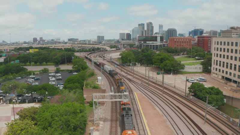 Aerial view of cargo train slowly riding through EBJ Union station. Backwards flying drone following railway corridor. Skyline with skyscrapers in background. Dallas, Texas, US