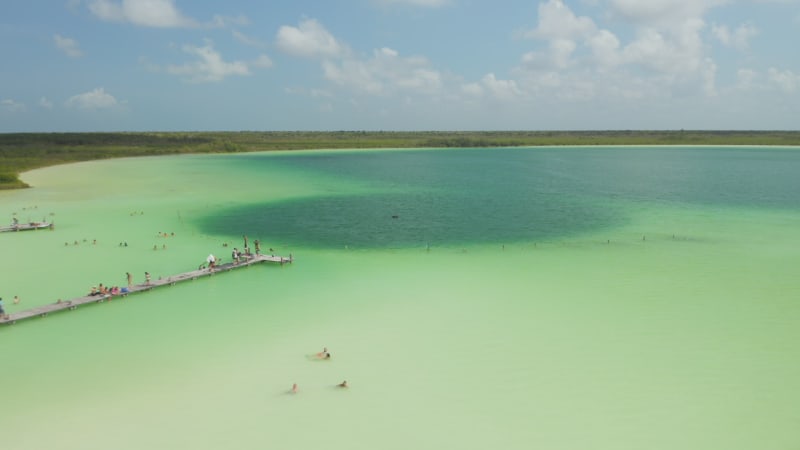 Backwards fly above amazing colourful lake in nature. People swimming and taking rest in sunny day. Kaan Luum lagoon, Tulum, Yucatan, Mexico