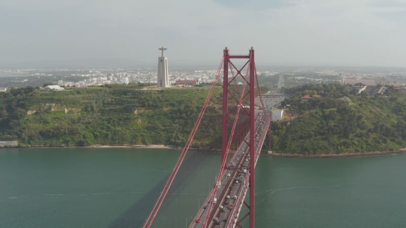 Aerial wide orbit of large tall monument of Santuario de Cristo Rei with lights and red Ponte 25 de Abril bridge across the sea near the coast of Lisbon, Portugal at night