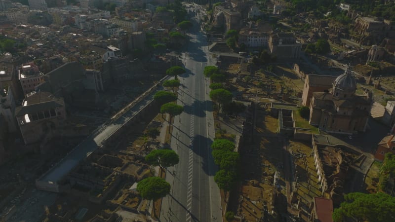 High angle view of road in city surrounded by remains of ancient sites. Tilt up reveal of cityscape with Colosseum amphitheatre in distance. Rome, Italy