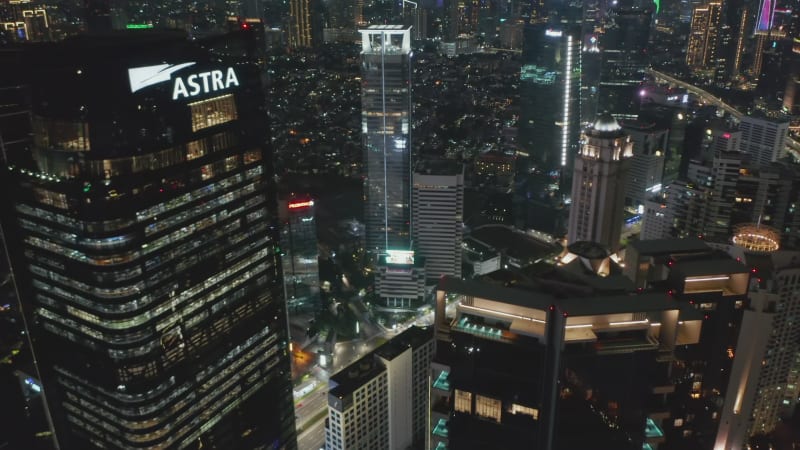 Close up fly by aerial shot of modern tall skyscrapers at night with busy car traffic below in Jakarta