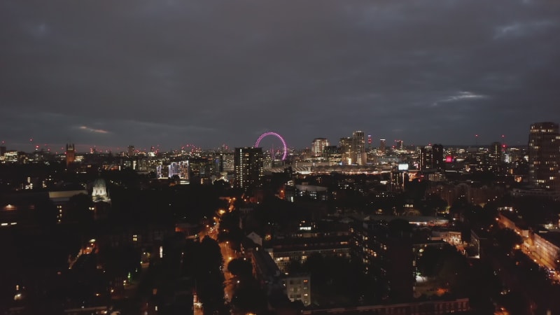 Descending shot of night cityscape. Urban neighbourhood at night. Illuminated London Eye in distance. London, UK