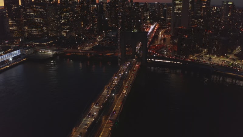 Fly above Brooklyn Bridge at night. Slowly moving vehicles on large bridge over river. Lighted windows of high rise buildings on waterfront. Manhattan, New York City, USA