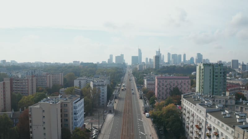 Backwards fly above wide and straight boulevard in city. Multilane road and tram track. Skyline with modern high rise office buildings. Warsaw, Poland