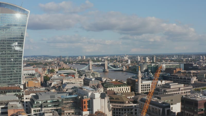 Fly above The City district. Glass facade on modern skyscraper reflecting sky with clouds and shining sun. Historic Tower Bridge over River Thames in background. London, UK