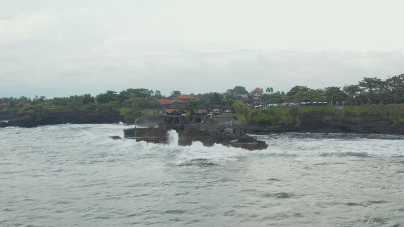 Low aerial shot approaching famous Tanah Lot temple tourist destination in Bali, Indonesia. Dangerous rough sea and strong waves crashing ashore small island on Bali coastline