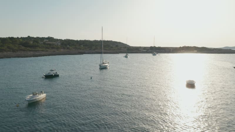 Aerial forward view of boats parked in a silent and calm sea surrounded by mountain and greenery at sunset in Ibiza in Spain