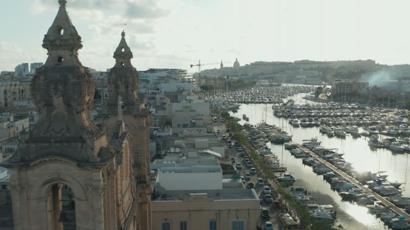 Small village in Malta revealing behind two Church Bell towers with boats in port on Sunny day, Aerial View