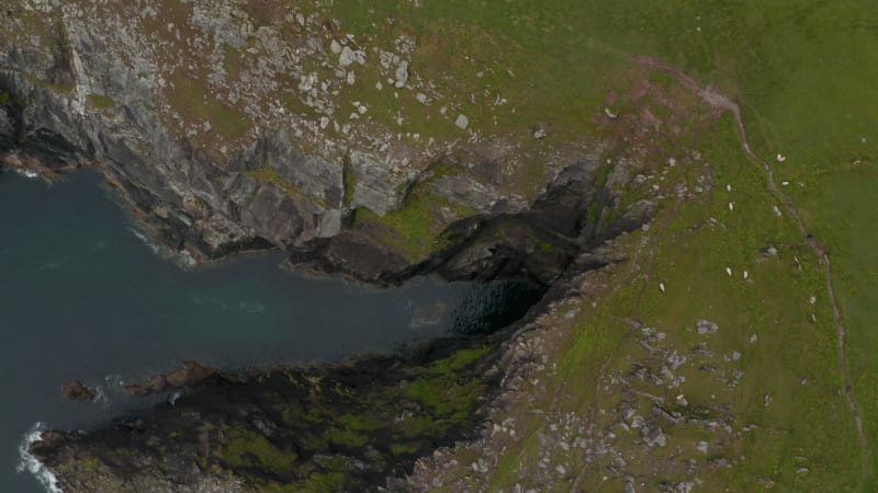 Aerial birds eye overhead top down view of rugged sea coast. Calm water surface under high rocky cliff. People walking on panoramic route along shore. Ireland