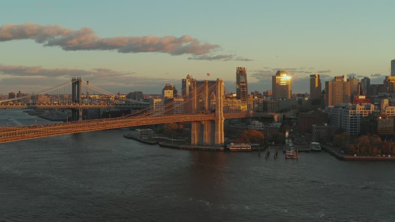 Large suspended bridge lit by setting sun. Cruise ship heading to Brooklyn Bridge. Buildings on waterfront. Brooklyn, New York City, USA