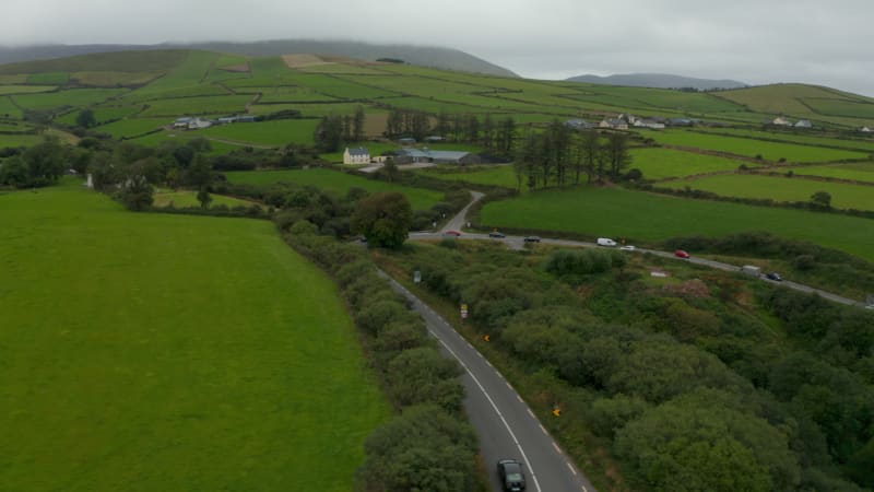 Aerial footage of green agricultural landscape. Vehicles passing through sharp curve on road. Hills shrouded in low clouds in distance. Ireland