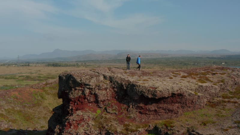 Orbit drone view birds eye two men standing cliff in Iceland looking panorama. Aerial view of two backpackers enjoying landscape and beauty in nature over hill