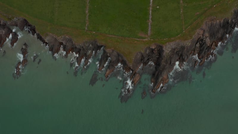 Aerial birds eye overhead top down view of waves crashing on jagged rocky sea coast. dark rocks rising above sand seabed. Ireland