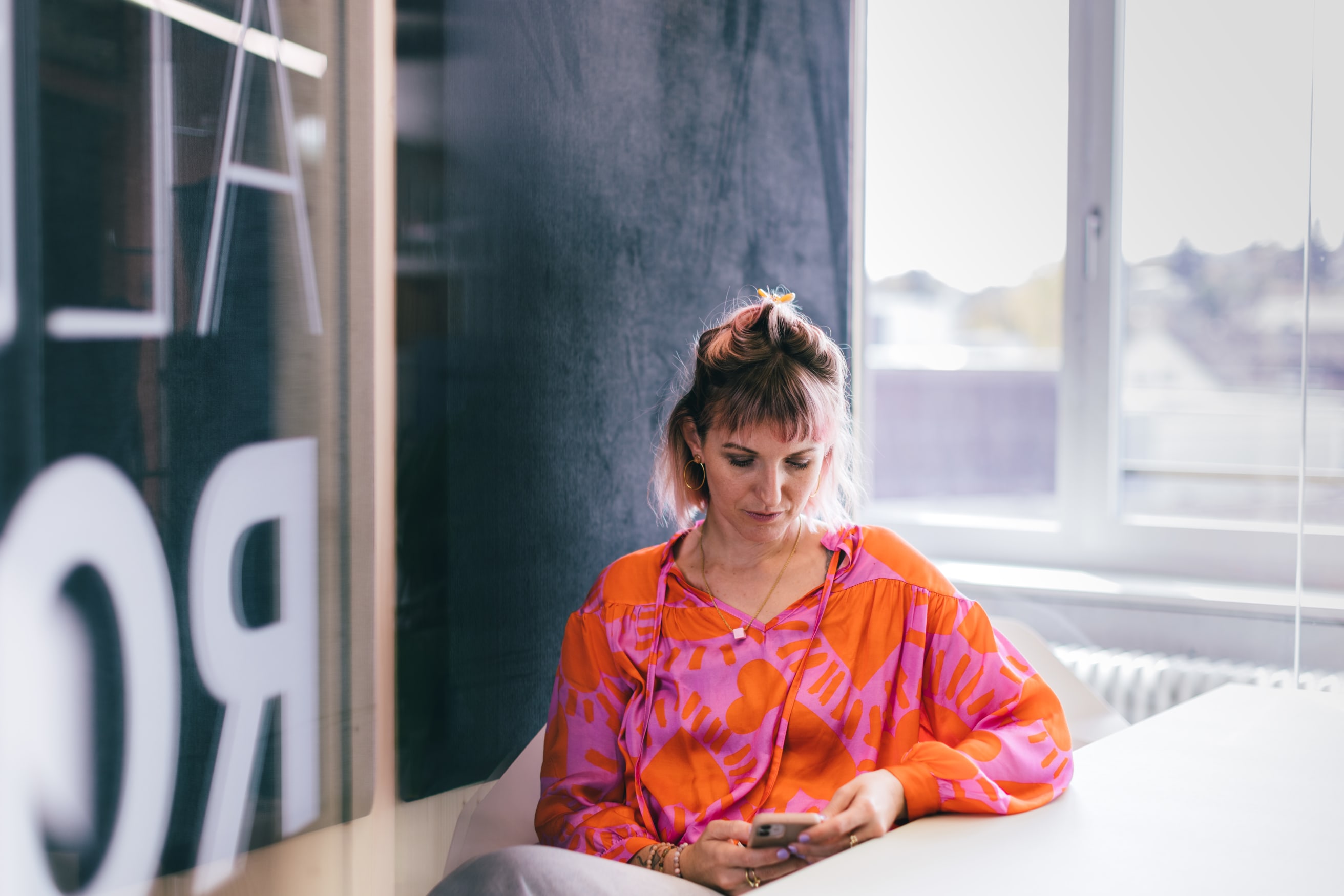The picture shows UX designer Natalie Kauer. She is sitting at a table and looking at a smartphone. She is wearing a colourful shirt.