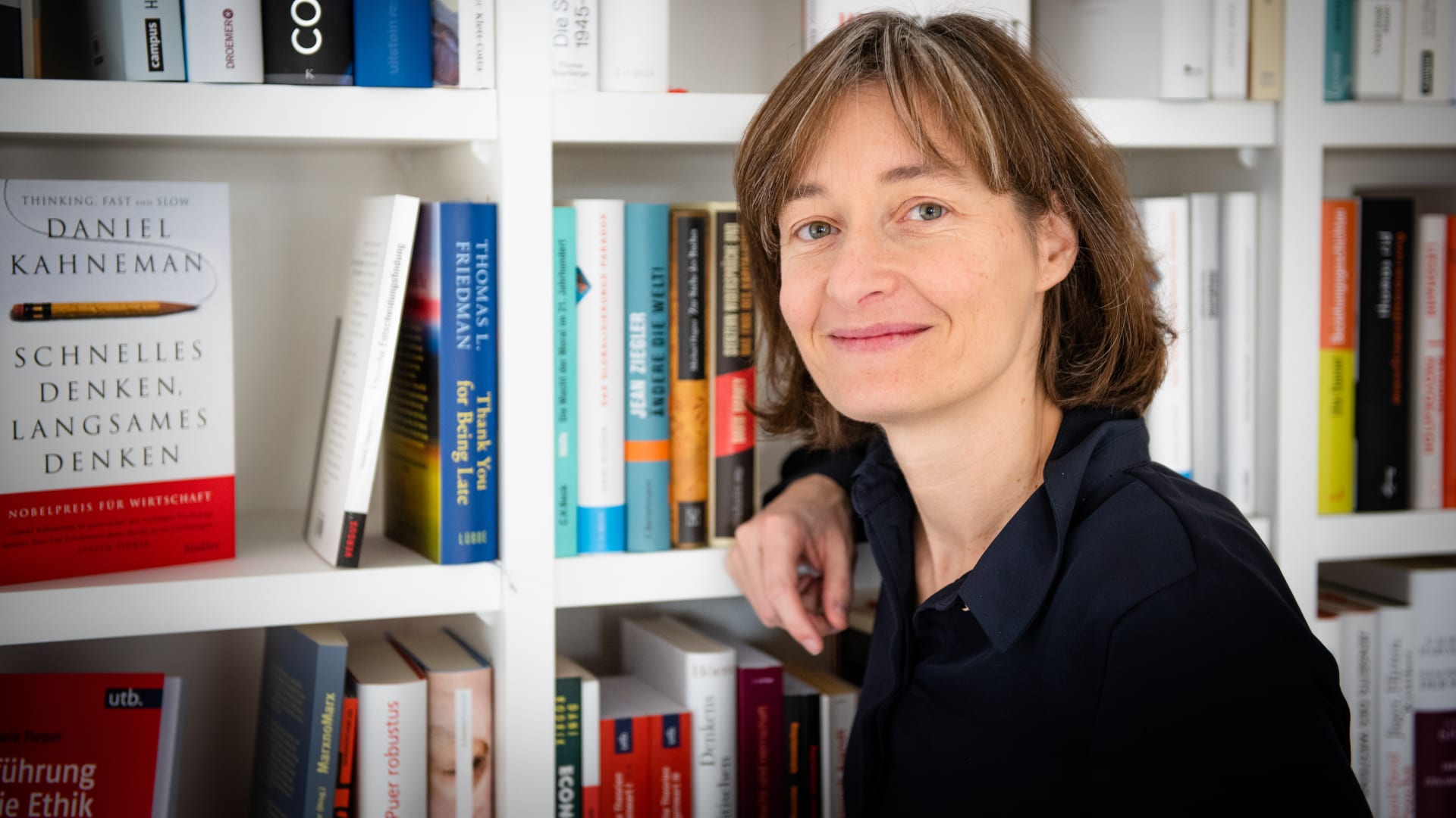 The picture shows Cornelia Diethelm in front of a full bookshelf.