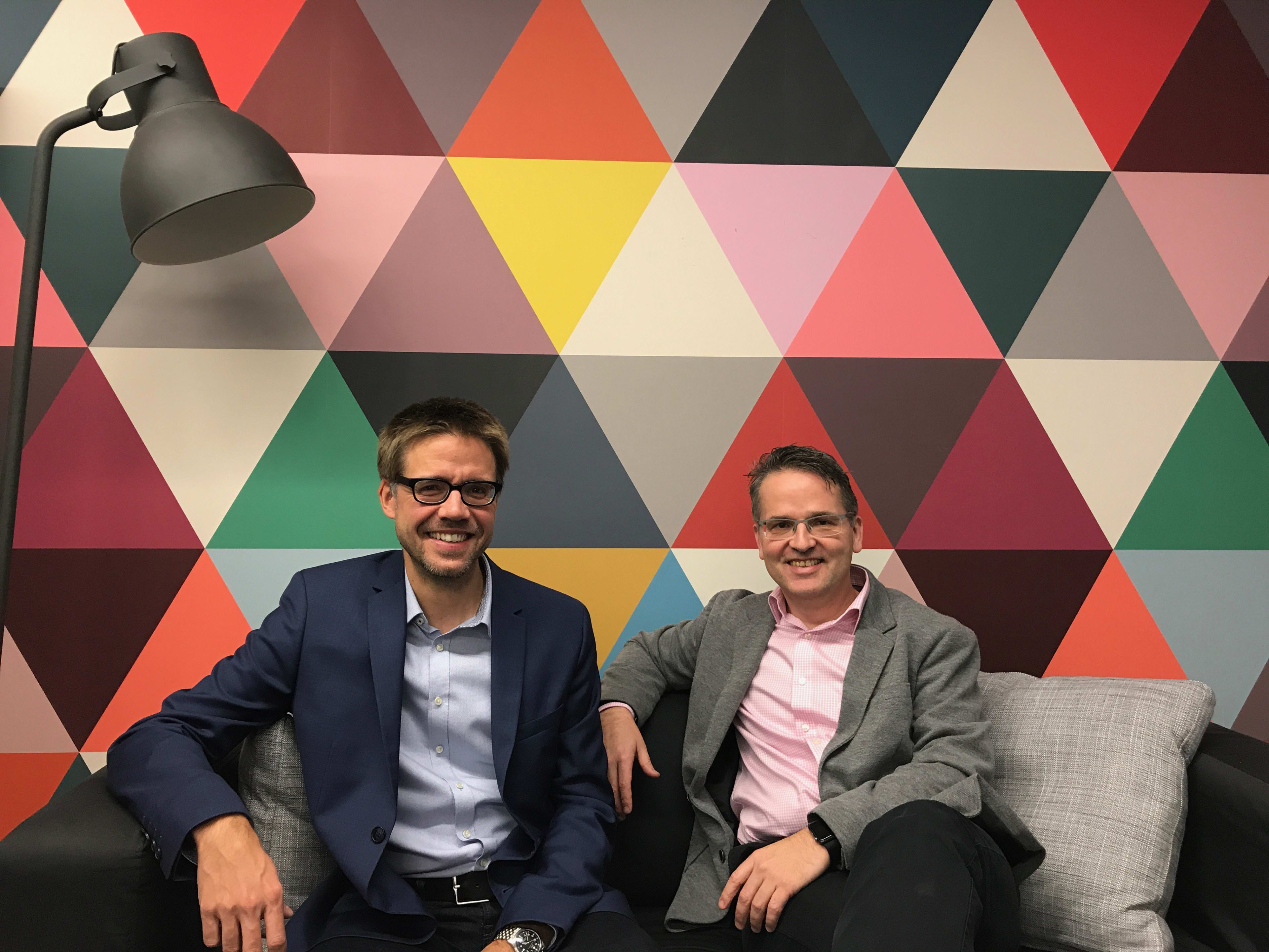 The picture shows the two Unic employees and Holacracy specialists Sandro Dönni and Ivo Bättig. They are sitting on a couch, smiling at the camera. Behind them you can see a brightly coloured wall of triangles.