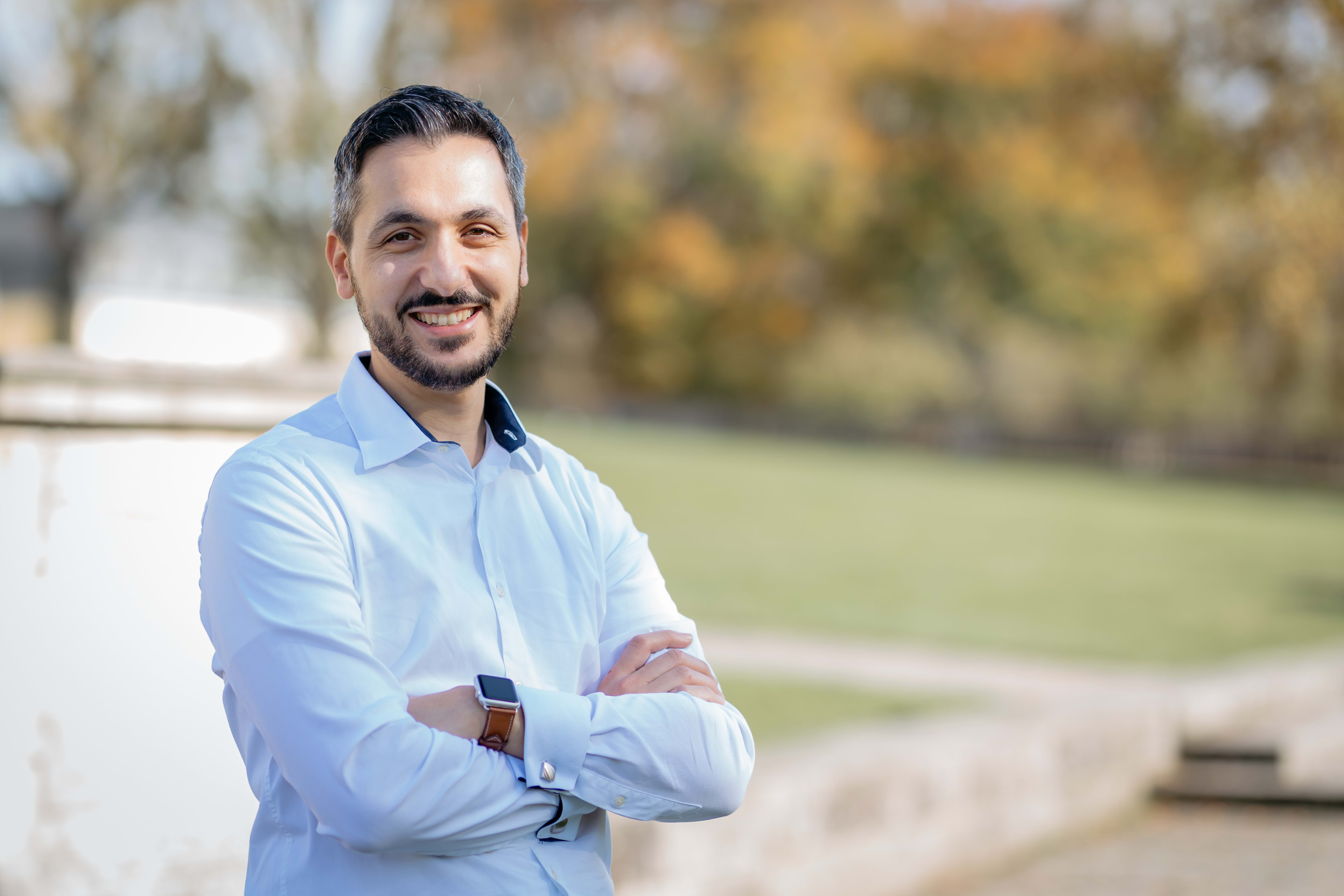 The picture shows a smiling man with a beard. He is wearing a light blue shirt and an Apple Watch with a brown strap. His arms are crossed.