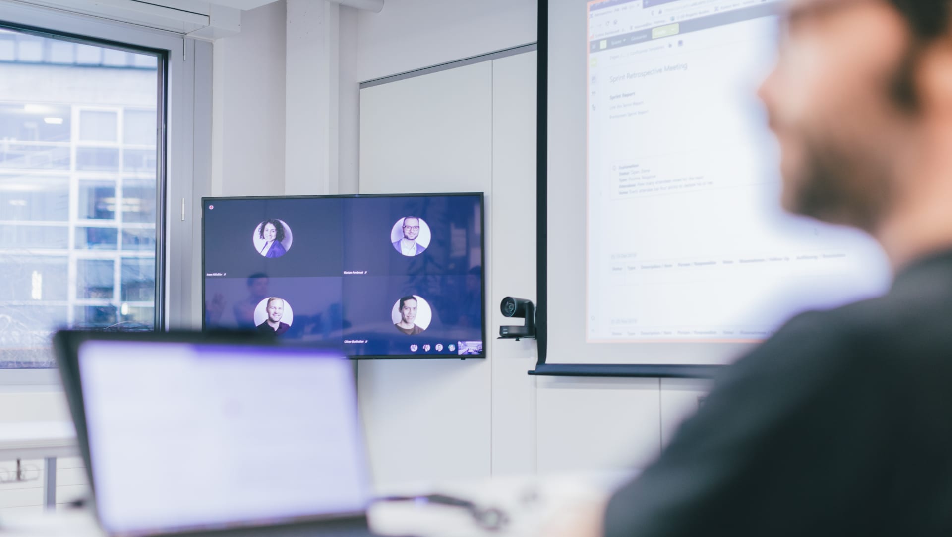 The picture shows a meeting in progress. There is at least one participant on site as well as participants who can be recognised on a TV set. At the same time, something is being presented on a projector board.