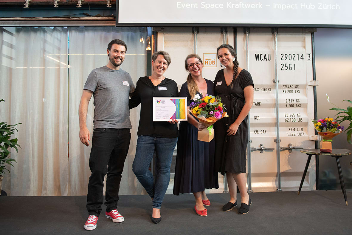 The picture shows 4 people smiling at the camera. One woman is holding a certificate with the LGBTI label, another is holding a bouquet of flowers.