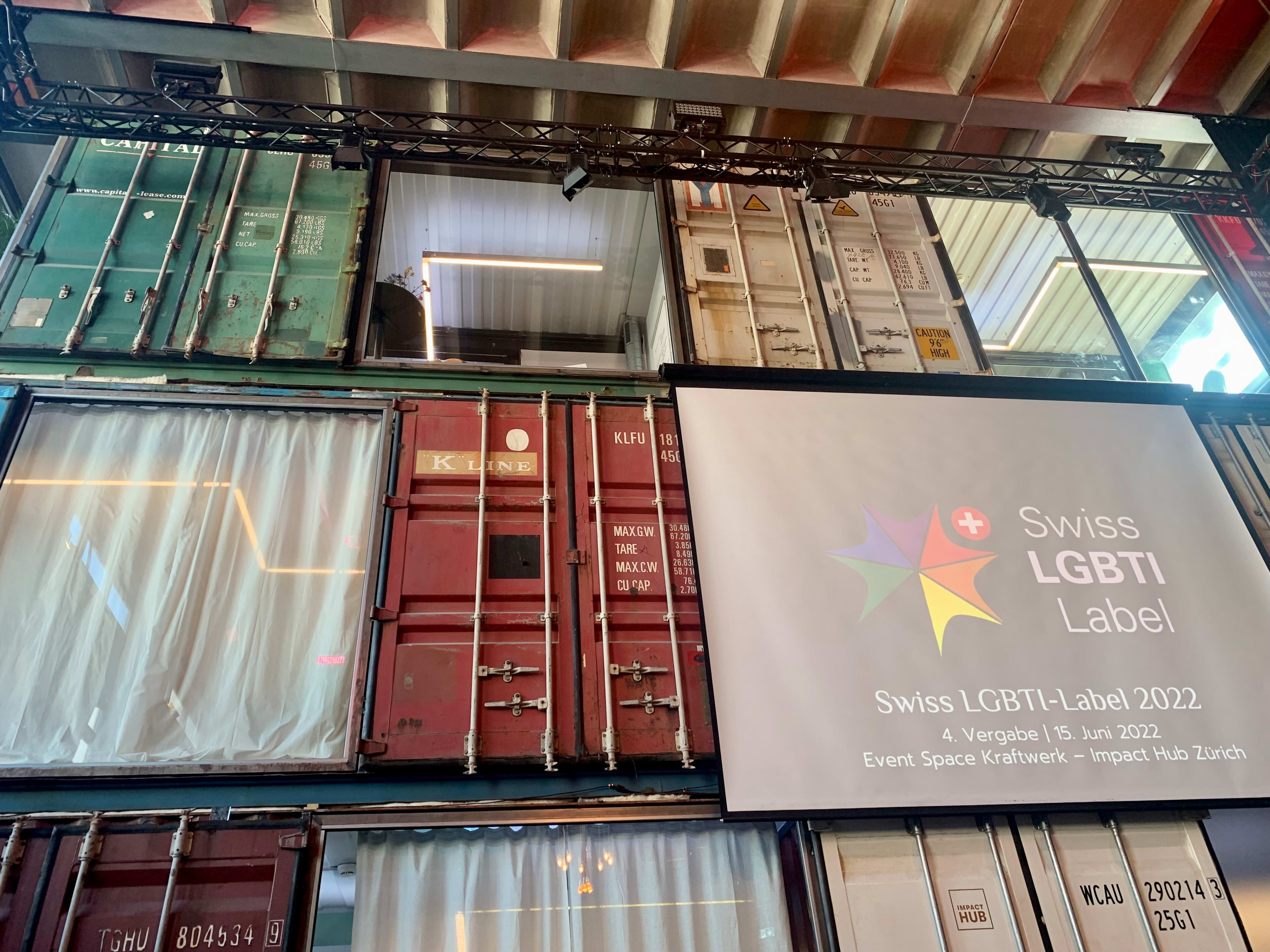 Das Bild zeigt gestapelte Container-Büros. Auf der rechten Bildseite ist das Plakat Swiss LGBTI Label zu sehen. Es handelt sich um eine Veranstaltung von 2022.