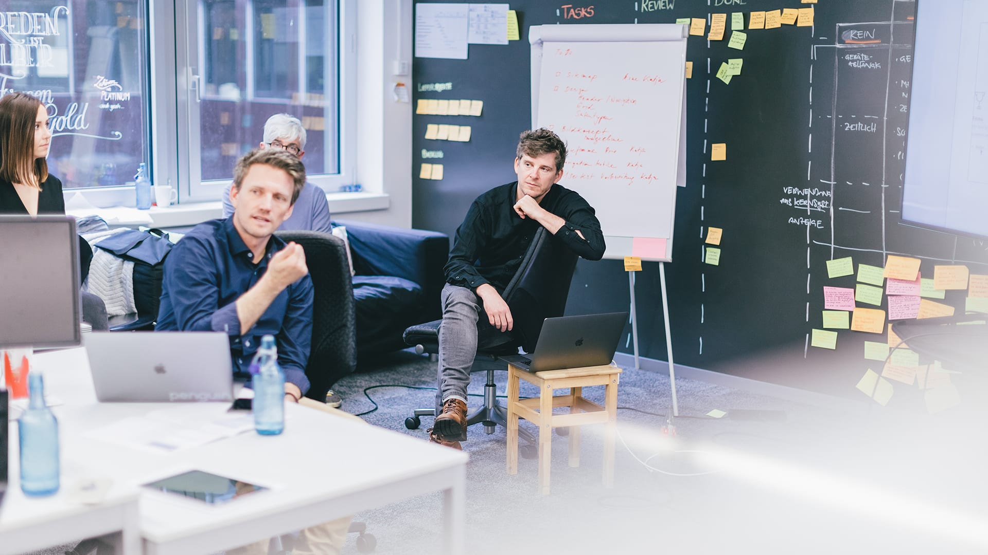 The picture shows several Unic employees discussing in a meeting room - in the background you can see a kind of Kanban board on a blackboard.