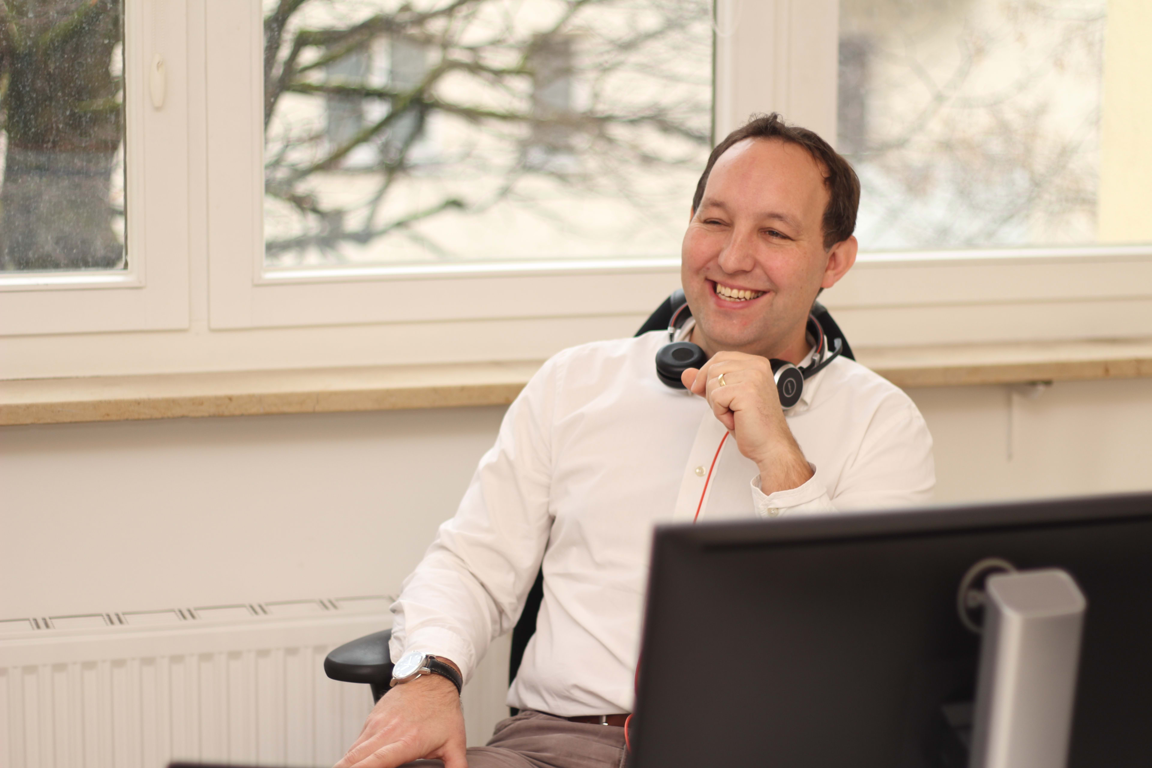 Das Bild zeigt den Senior Consultant Peter Köllner von Unic. Er sitzt am Schreibtisch und lacht in den Raum hinein. Man erkennt das Headset um seinen Hals. Im Vordergrund steht ein Monitor.