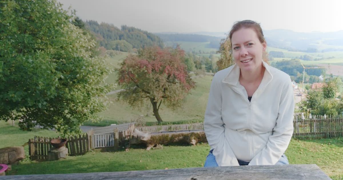 Das Bild zeigt eine Frau, sitzend am Holztisch. Im Hintergrund erkennt man eine eingezäunte Rasenfläche und dahinter die weite Flur mit Wiesen und Bäumen.