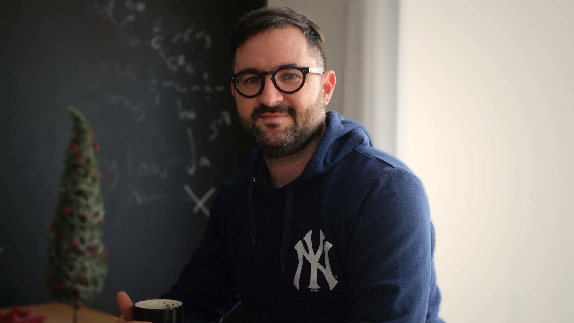 Das Bild zeigt einen Mann mit Brille und Bart. Er trägt einen Pulli der New York Yankees. Vor ihm steht eine Tasse.