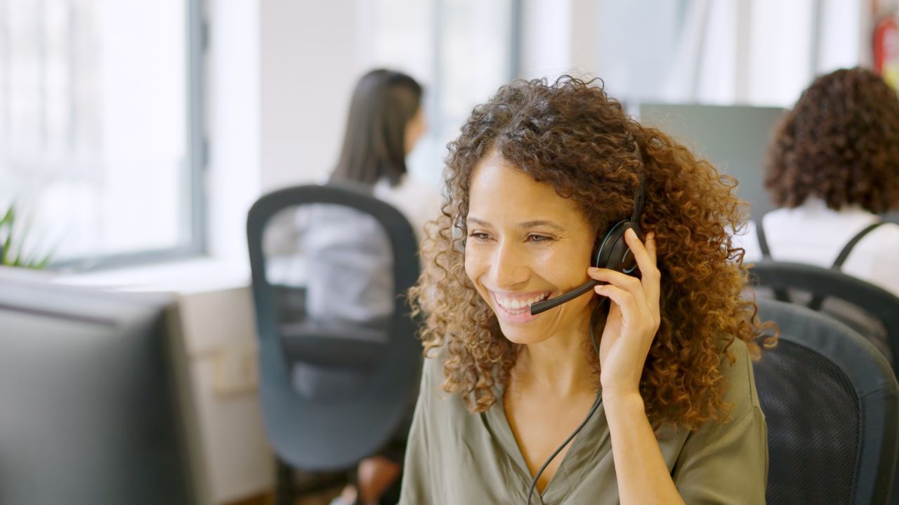 A woman wearing a headset smiles while working at a computer in an office—perfectly illustrating what is an answering service; other people are visible working in the background.