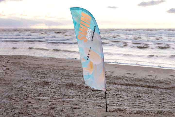 Zastava za plažu sa šarenim dizajnom i natpisom "SWIM" vijori se na pješčanoj plaži ispred valovitog mora, oblačnog neba u pozadini.