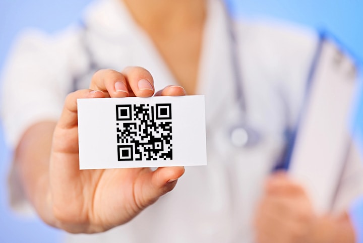Person in doctor's coat holding business card with large QR code in hand, blurred background showing medical documents, symbol of digital networking in healthcare.