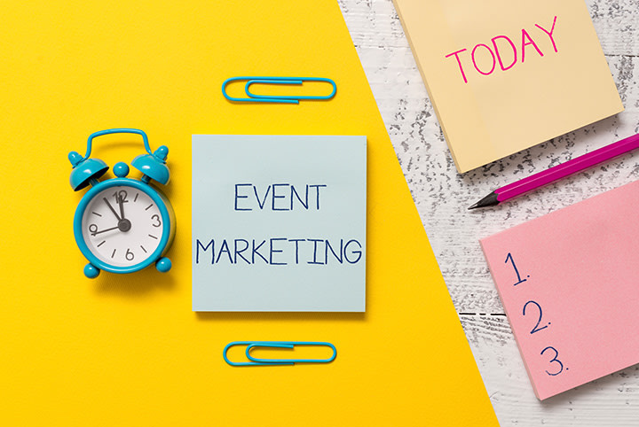 Blue alarm clock, sticky notes with "Event Marketing", "TODAY" and list on yellow table. Symbol for time management and event marketing.