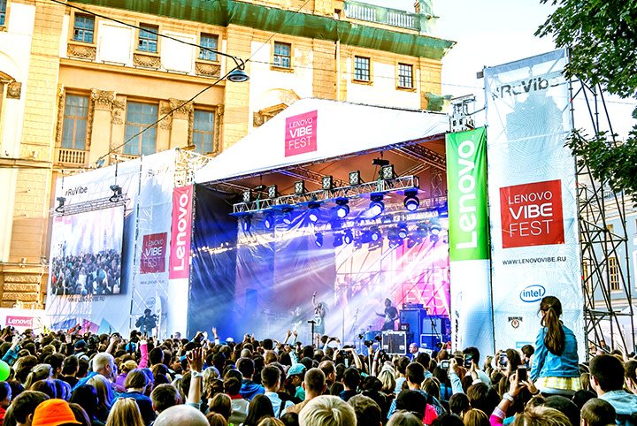 Open-Air-Konzert beim Lenovo Vibe Fest mit großer Menschenmenge vor der Bühne in urbaner Umgebung, bunten Lichtern und markanten Lenovo-Werbeschildern.