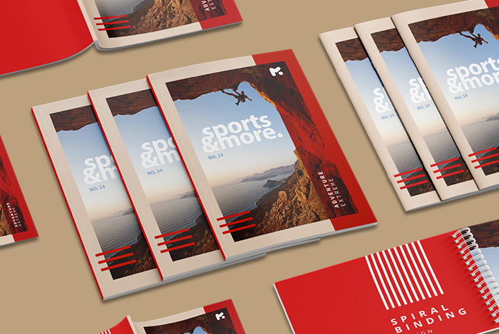 Kelios raudonos ir baltos spalvos brošiūros, pavadintos "Sportas ir daugiau", alpinisto ant uolos veido su vaizdu į jūrą, atvaizdas, išsidėstęs ant stalo.
