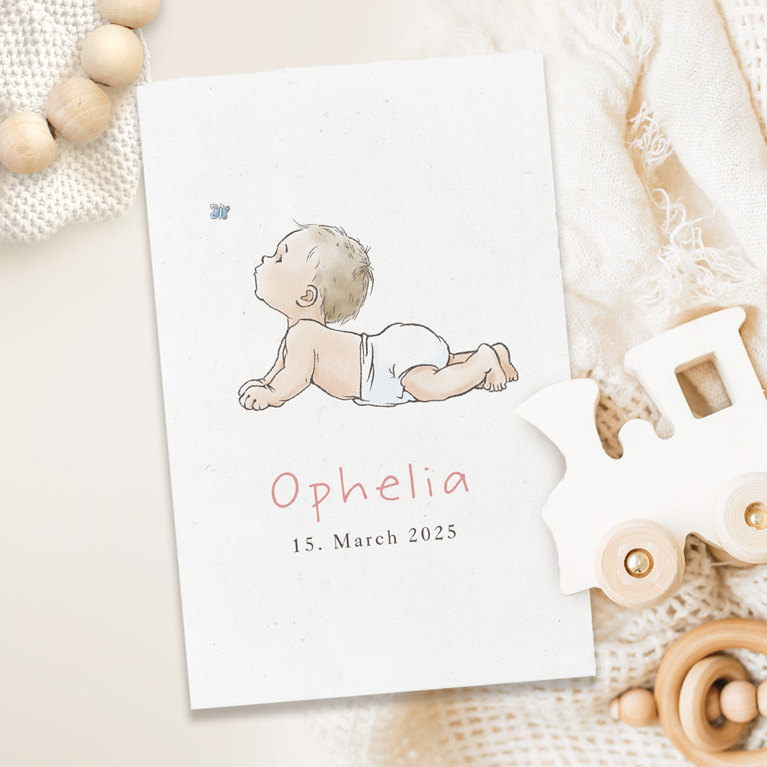 Anlasskachel zum Thema Geburt: Klappkarte mit gezeichneter Babyillustration, umgeben von Holzspielzeug und gestrickter Decke auf hellem Hintergrund.